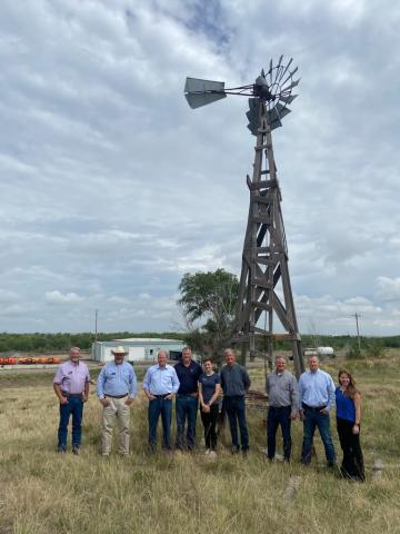 Governor Polis standing with local leaders in a field in front of a tall wooden wind mill.